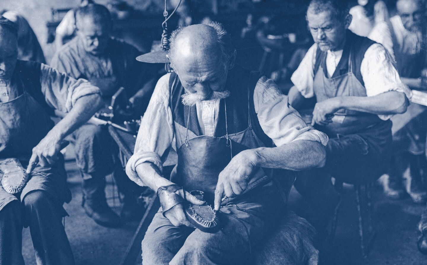 Foto von 1899, mehrere Schuhmacher bearbeiten jeweils einen Schuh, der in ihrem Schoß liegt.