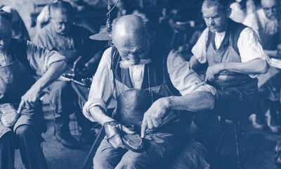Foto von 1899, mehrere Schuhmacher bearbeiten jeweils einen Schuh, der in ihrem Schoß liegt.